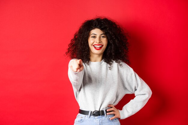 Smiling cheerful woman with curly hairstyle pointing fingers at camera inviting you choosing someone...