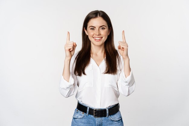 Smiling confident pretty woman, pointing fingers up, showing logo banner, announcement, standing in white blouse against studio background.