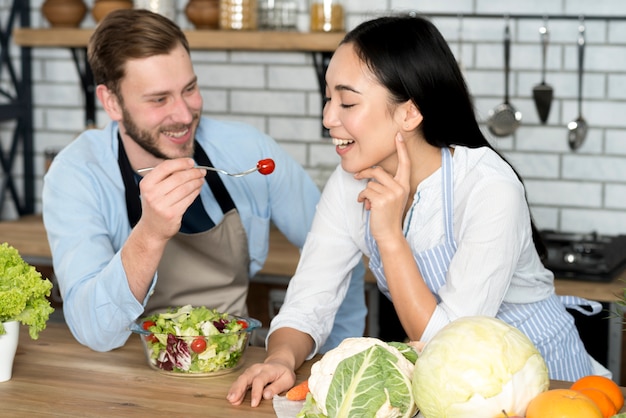 Free photo smiling couple feeding healthy salad in kitchen wearing apron