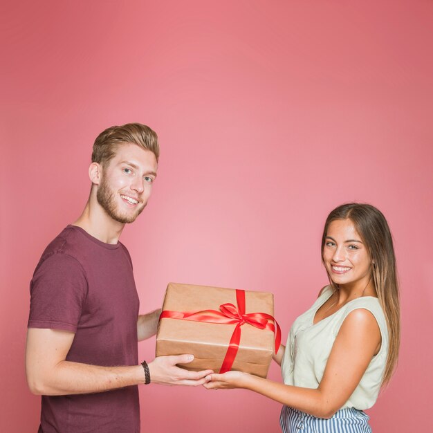 Smiling couple holding gift box with red satin ribbon against pink background