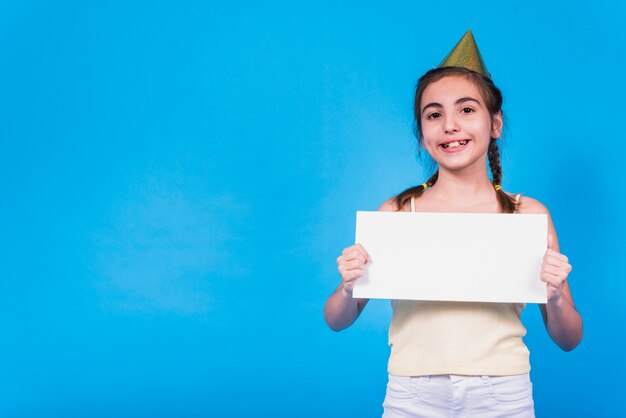 Smiling cute girl wearing party hat holding blank card in hand in front of colored wallpaper