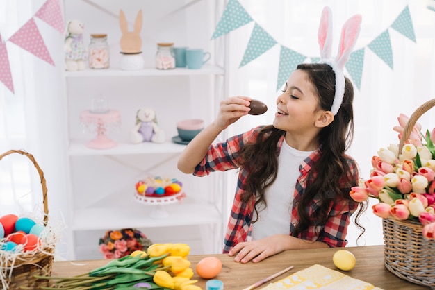 Smiling cute girl with bunny ears over her head eating chocolate easter egg