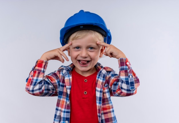 Free photo a smiling cute little boy with blonde hair wearing checked shirt in blue helmet pointing at his head with index fingers while looking on a white wall