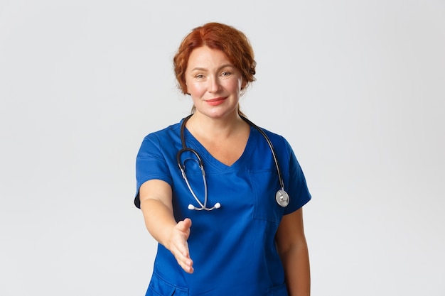 Smiling cute middle-aged female nurse, doctor in blue scrubs looking friendly, extend hand for handshake, introduce herself, 