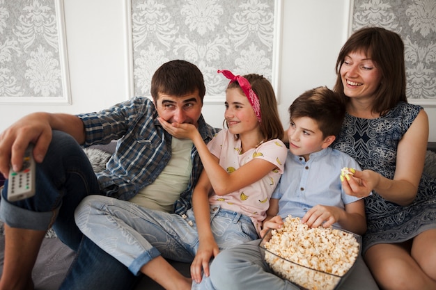 Smiling daughter sitting on sofa with family and feeding popcorn to her father