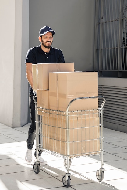 Free photo smiling delivery walking on pavement with trolley full of cardboard boxes