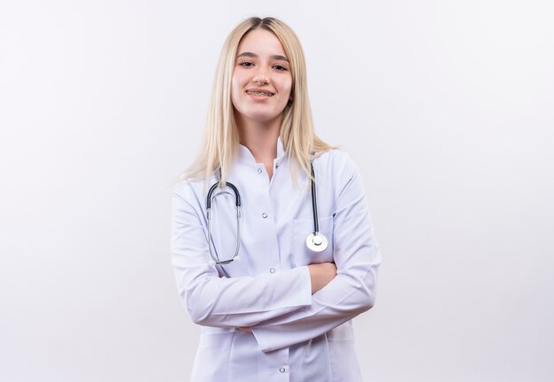 Smiling doctor young blonde girl wearing stethoscope and medical gown in dental brace crossing hands on isolated white background