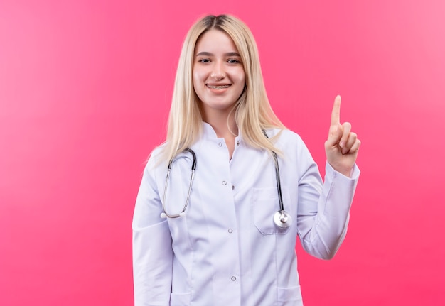 Smiling doctor young blonde girl wearing stethoscope in medical gown and dental brace points to up on isolated pink background