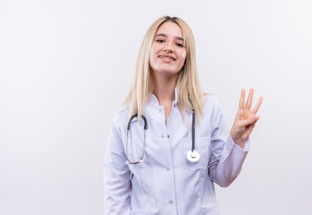 Smiling doctor young blonde girl wearing stethoscope and medical gown in dental brace showing three on isolated white background