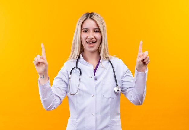 Smiling doctor young girl wearing stethoscope in medical gown and dental brace points to up on isolated yellow background