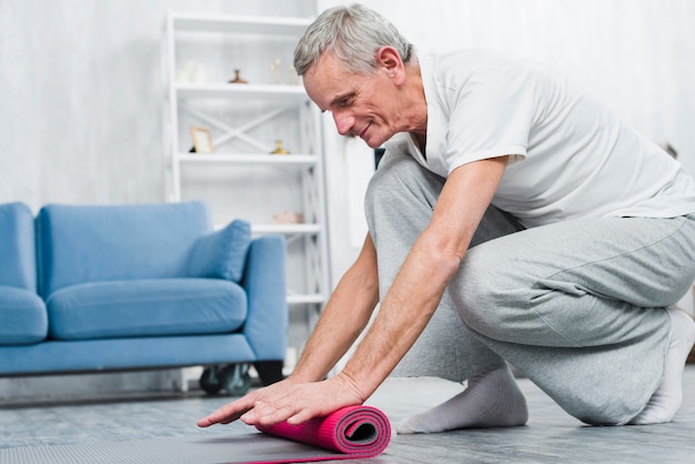 Smiling elder man rolling yoga mat after yoga in home