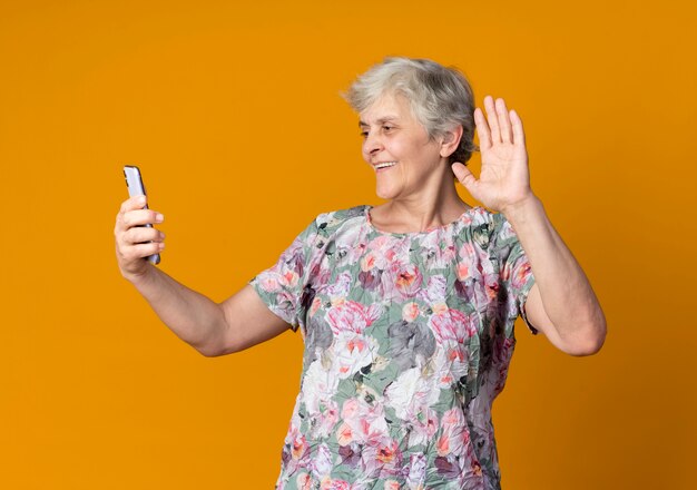 Smiling elderly woman raises hand holding and looking at phone isolated on orange wall