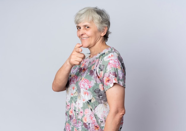 Smiling elderly woman stands sideways pointing isolated on white wall