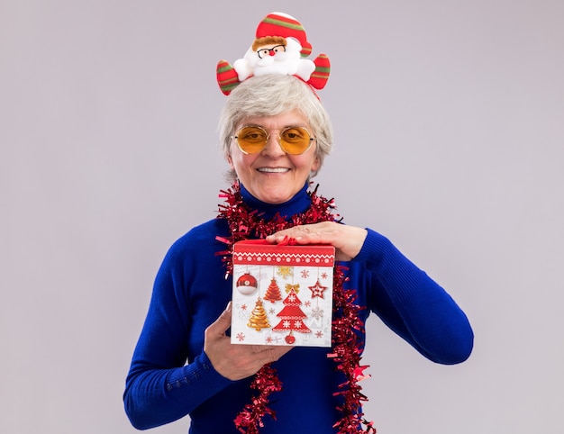 Smiling elderly woman in sun glasses with santa headband and garland around neck holds christmas gift box isolated on white wall with copy space