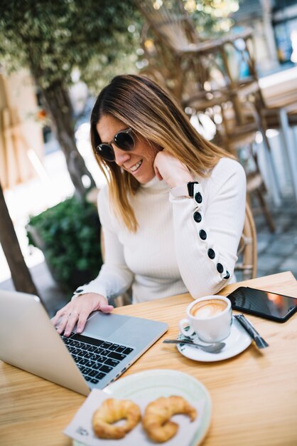 Smiling elegant young woman using laptop at table with drink and croissants in street cafe