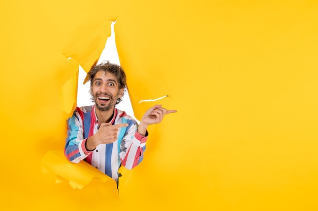 Smiling and emotional young man poses in torn yellow paper hole background and pointing something