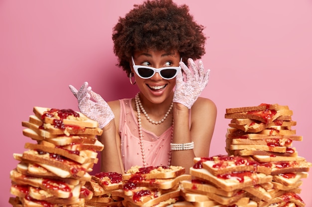 Free photo smiling ethnic lady looks aside and keeps hand on sunglasses, being in good mood, giggles positively on party, wears stylish clothes, poses over rosy wall, many delicious sandwiches around