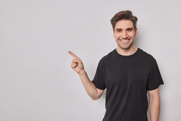 Smiling European man with glad expression points at blank copy space shows promotion offer demonstrates advertisement wears casual black t shirt isolated over white background. Look at this.