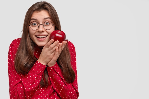 Smiling European woman with pleased expression, carries red apple, dressed in fashionable clothes, round spectacles, enjoys eating fruit