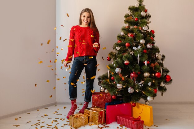 Smiling excited pretty woman in red sweater sitting at home at Christmas tree throwing golden confetti surrounded with presents and gift boxes