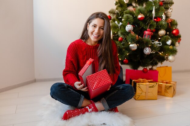 Smiling excited pretty woman in red sweater sitting at home at Christmas tree unpacking presents and gift boxes