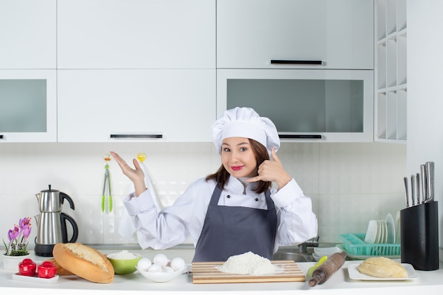 Smiling female chef in uniform standing behind the table with cutting board bread vegetables making call me gesture in the white kitchen