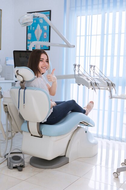 Smiling female patient sitting on chair showing thumb-up in dental clinic