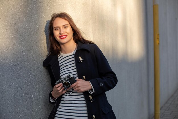 Smiling female photographer in jacket standing in front of wall ready to make new photo. Adorable young brunette woman in trendy outfit posing on concrete wall background with camera