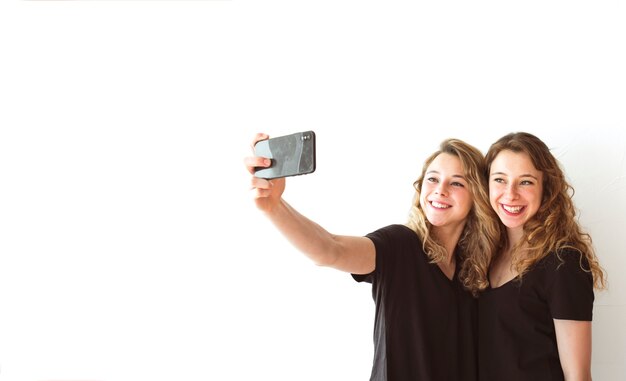 Smiling female sister taking selfie on cellphone against white backdrop