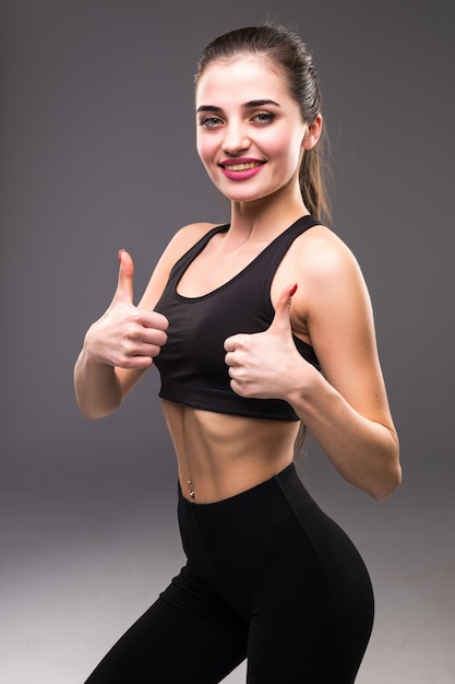 Smiling fitness woman showing thumb up sign over gray wall