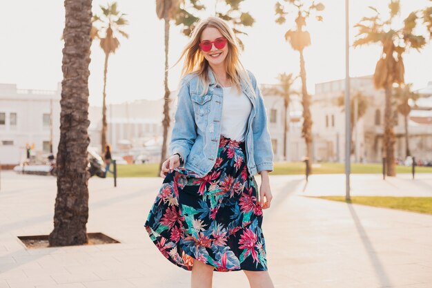 smiling flirting woman walking in city street in stylish printed skirt and denim oversize jacket wearing pink sunglasses