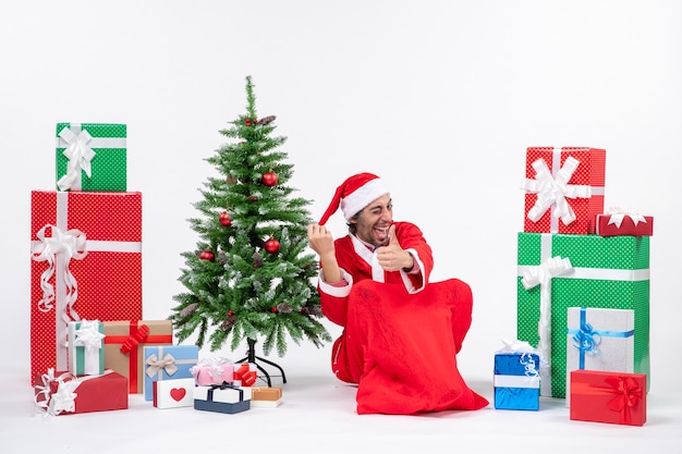 Free photo smiling funny young man dressed as santa claus with gifts and decorated christmas tree sitting on the ground making ok gesture on white background