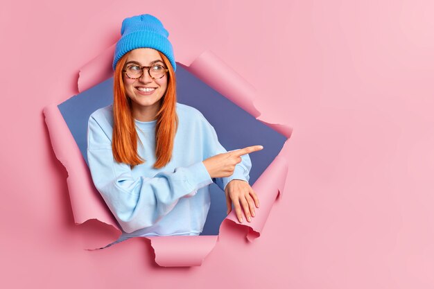 Smiling ginger woman feels optimistic pointing at copy space, wears blue hat glasses and jumper breaks through paper