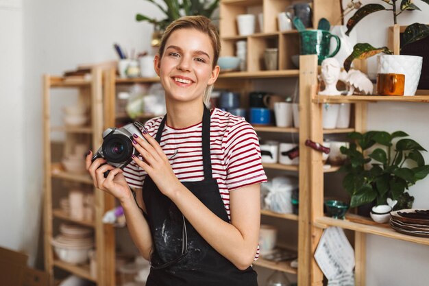 Smiling girl in black apron and striped T-shirt holding camera in hands happily looking in camera at pottery studio