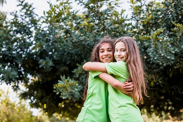 Smiling girl embracing each other against green tree