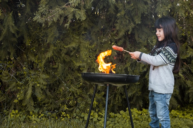 Free photo smiling girl grilling sausages on portable barbecue at outdoors