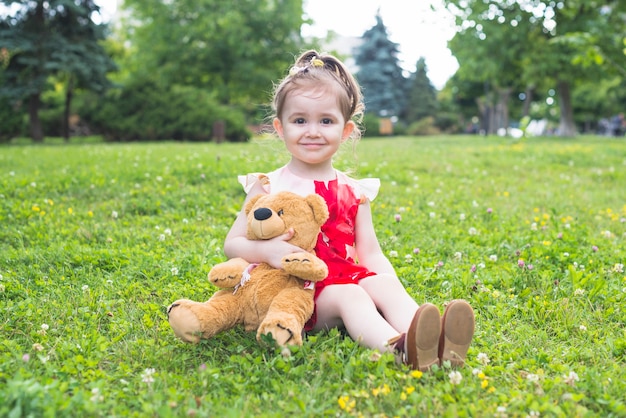 Free photo smiling girl holding teddy bear sitting on the green grass