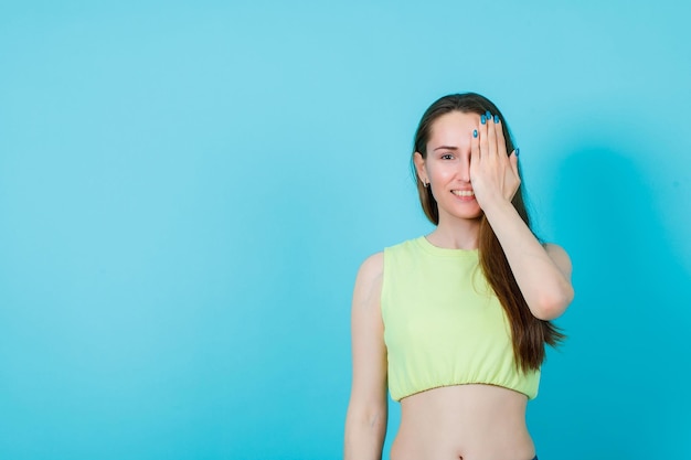 Smiling girl is coverig half face with hand on blue background