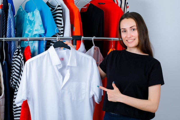 Smiling girl is holding shirt and showing it with forefinger on clothes background