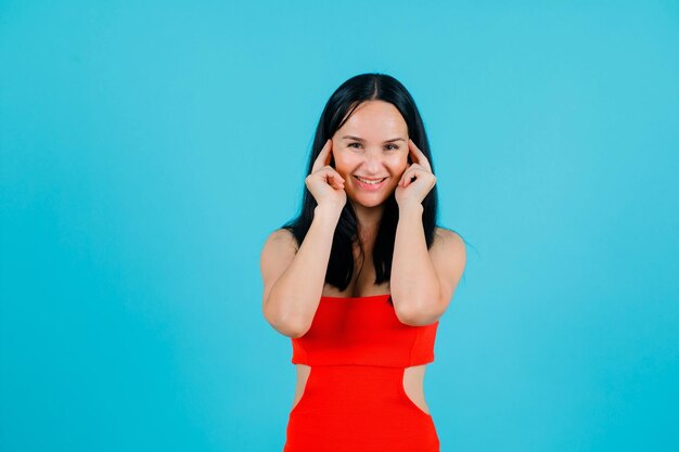 Smiling girl is looking at camera by holding forefinger on ears on blue background