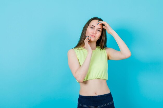 Smiling girl is looking at camera by holding hands on face on blue background