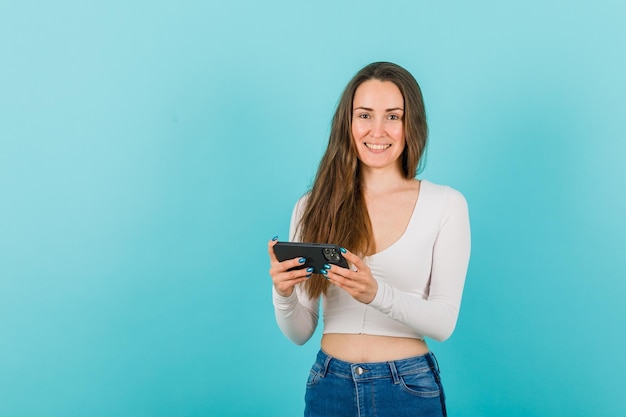 Smiling girl is looking at camera by holding smartphone on blue background