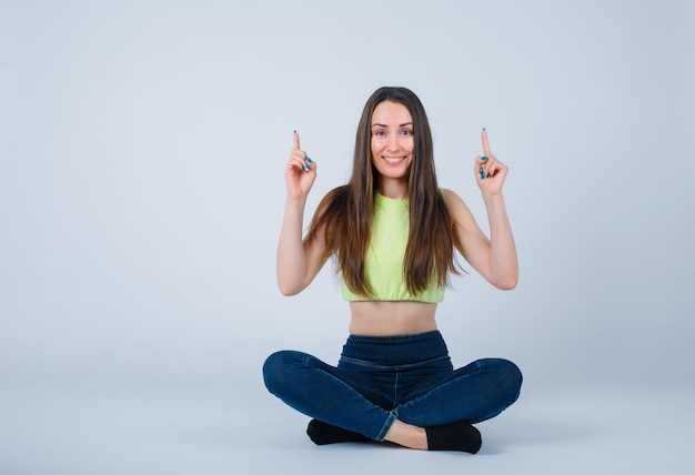 Smiling girl is pointing up with forefingers by sitting on flooor on white background