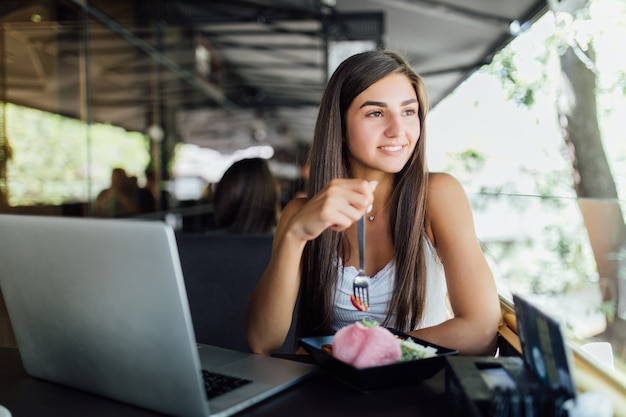 Smiling girl is sitting in cafe and works on her homework on laptop in daytime