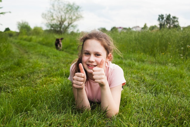 Free Photo smiling girl lying on green grass and showing thumb up gesture in park