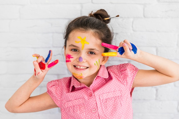 Smiling girl making peace gesture with painted fingers and face against white brick wall