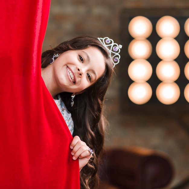 Free Photo smiling girl peeking behind the red curtain at backstage