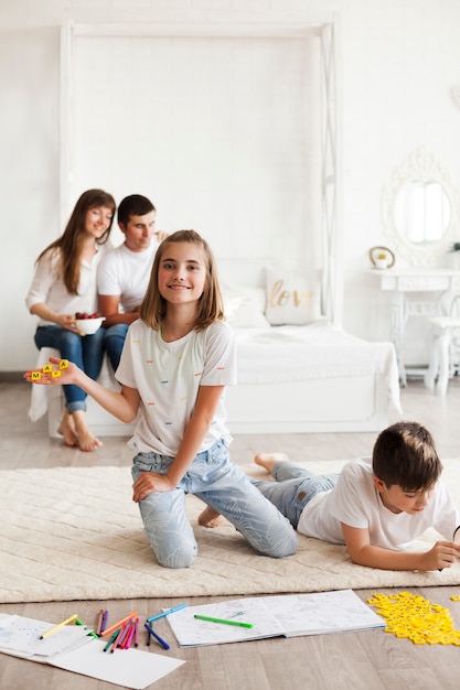 Smiling girl showing block of scrabble game letter while her parents sitting on bed