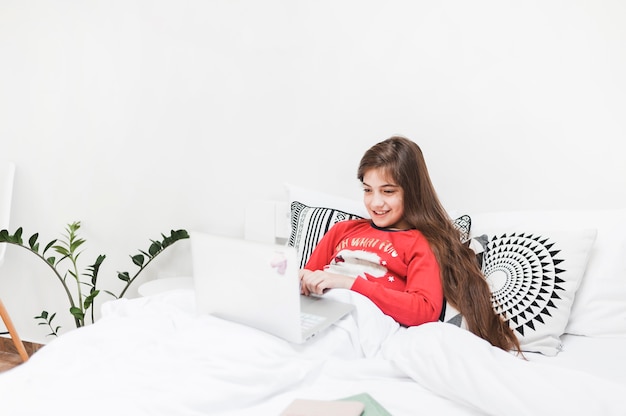 Smiling girl sitting on bed using laptop in bedroom