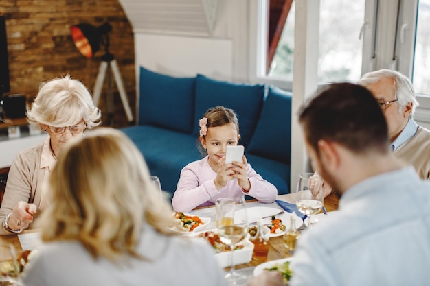 Free photo smiling girl sitting at dining table with her parents and grandparents and using cell phone